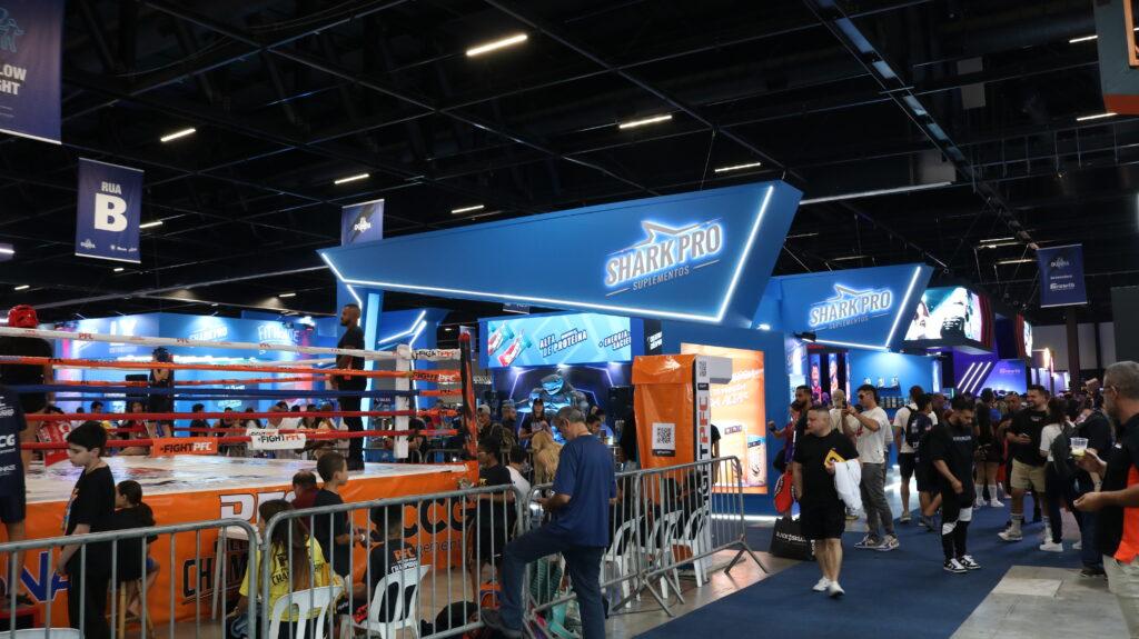 Multidão circula pela feira Mr. Olympia, com o ringue de boxe à esquerda e o estande da Shark Pro, cliente da LabelColor, visível ao fundo, contribuindo para a energia vibrante do evento.