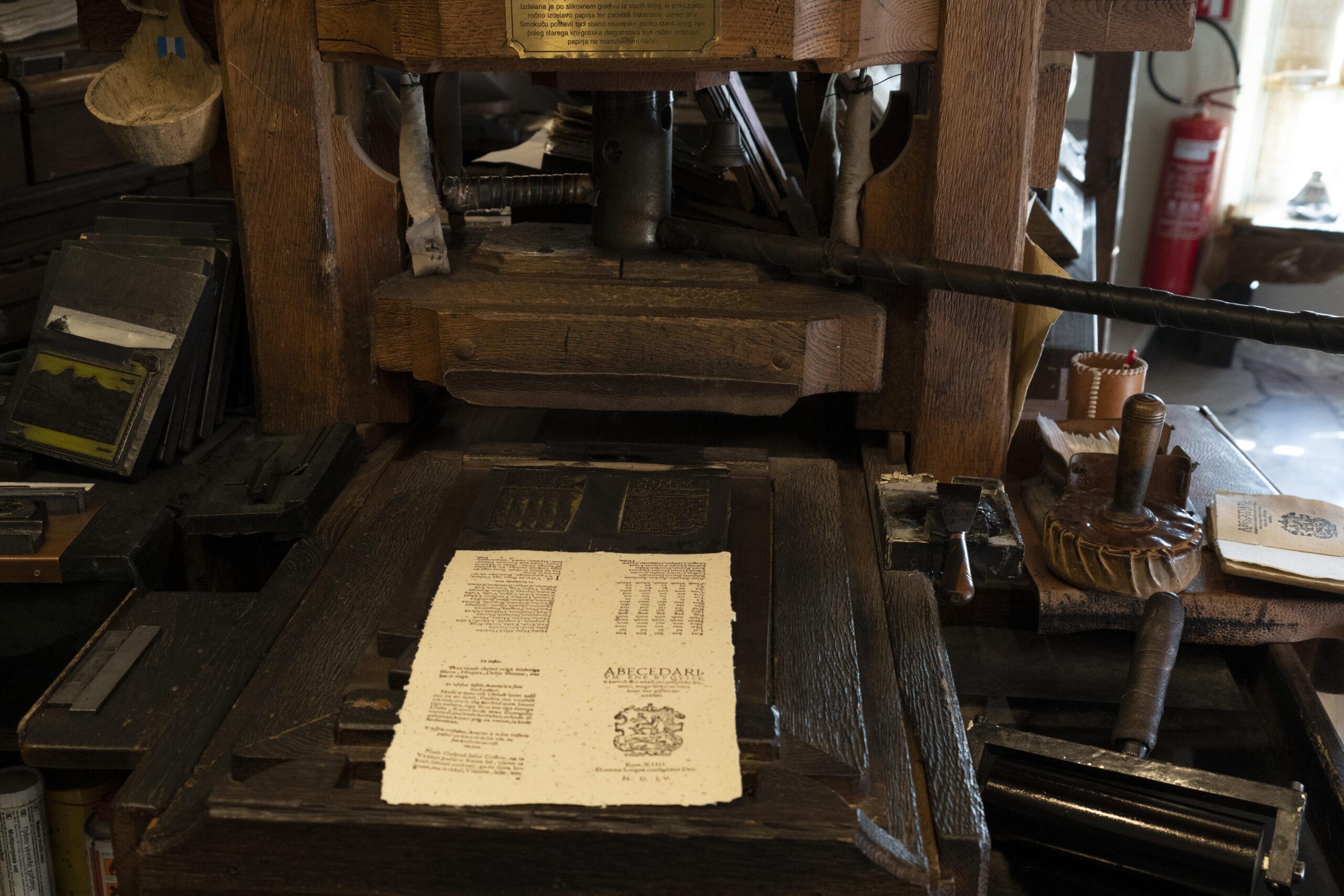 Fotografia de uma antiga prensa tipográfica de madeira, semelhante às usadas na época de Johannes Gutenberg. No centro da imagem, há uma folha impressa com texto e um brasão, posicionada sobre a matriz tipográfica. Ao redor, diversos instrumentos de impressão, como rolos de tinta, tipos móveis e ferramentas de entintagem, estão organizados em uma bancada de madeira. A cena remete ao processo tradicional de impressão, destacando a evolução da gráfica ao longo dos séculos.