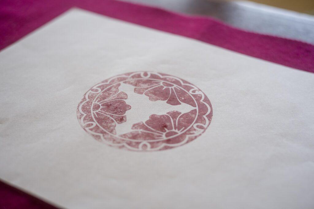 Impressão do carimbo em tinta vermelha sobre uma folha de papel branco. O desenho apresenta um padrão floral circular com detalhes ornamentais.